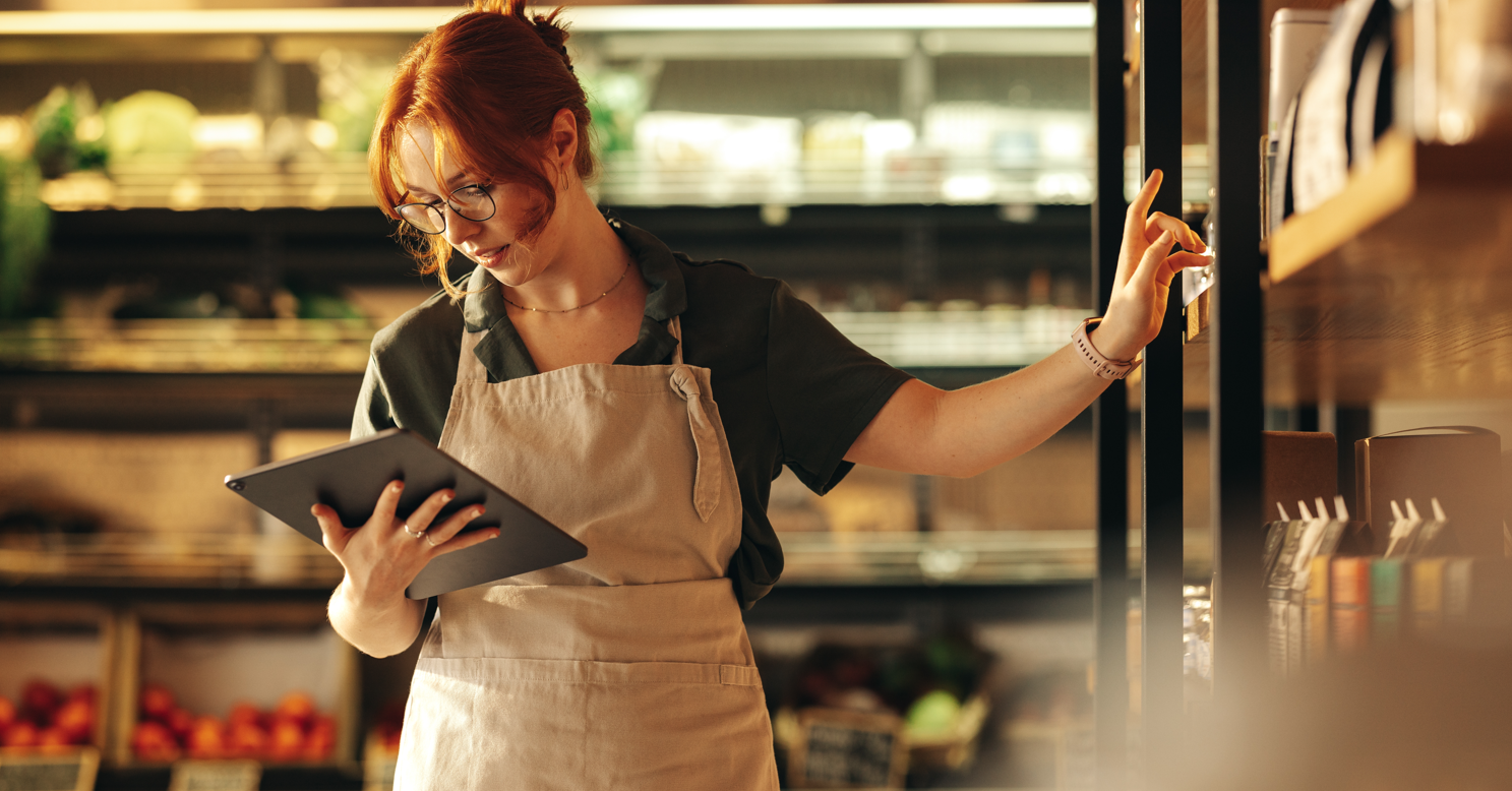 Woman with red hair, working at a grocery store, looking at a tablet in her hand