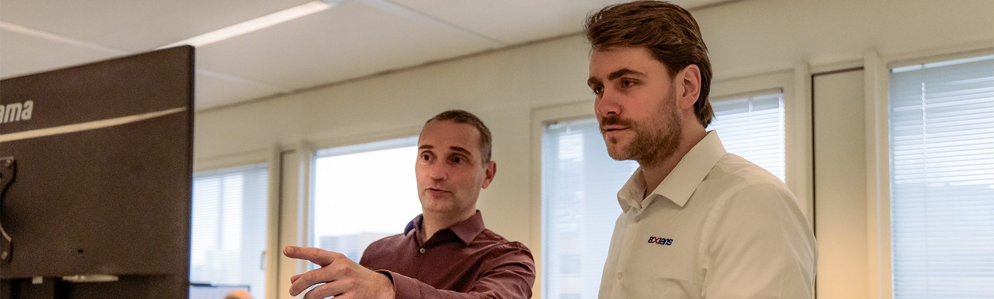 De IT-directeur van Mosaic World bespreekt een workflow met een Axians-consultant op kantoor. Ze kijken samen naar een scherm tijdens de opnames van de referentiecase over Exact Synergy.