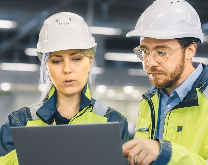 Industriële IT-professionals in veiligheidskleding werken samen in een fabriek of werkplaats. Ze bekijken gegevens op hun devices