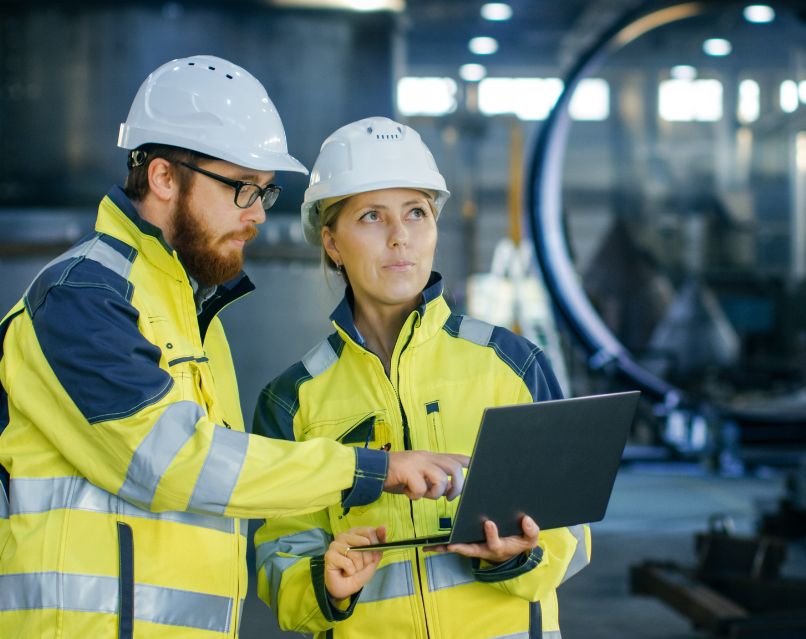 Industriële IT-professionals in veiligheidskleding werken samen in een fabriek of werkplaats. Ze bekijken gegevens op hun laptop